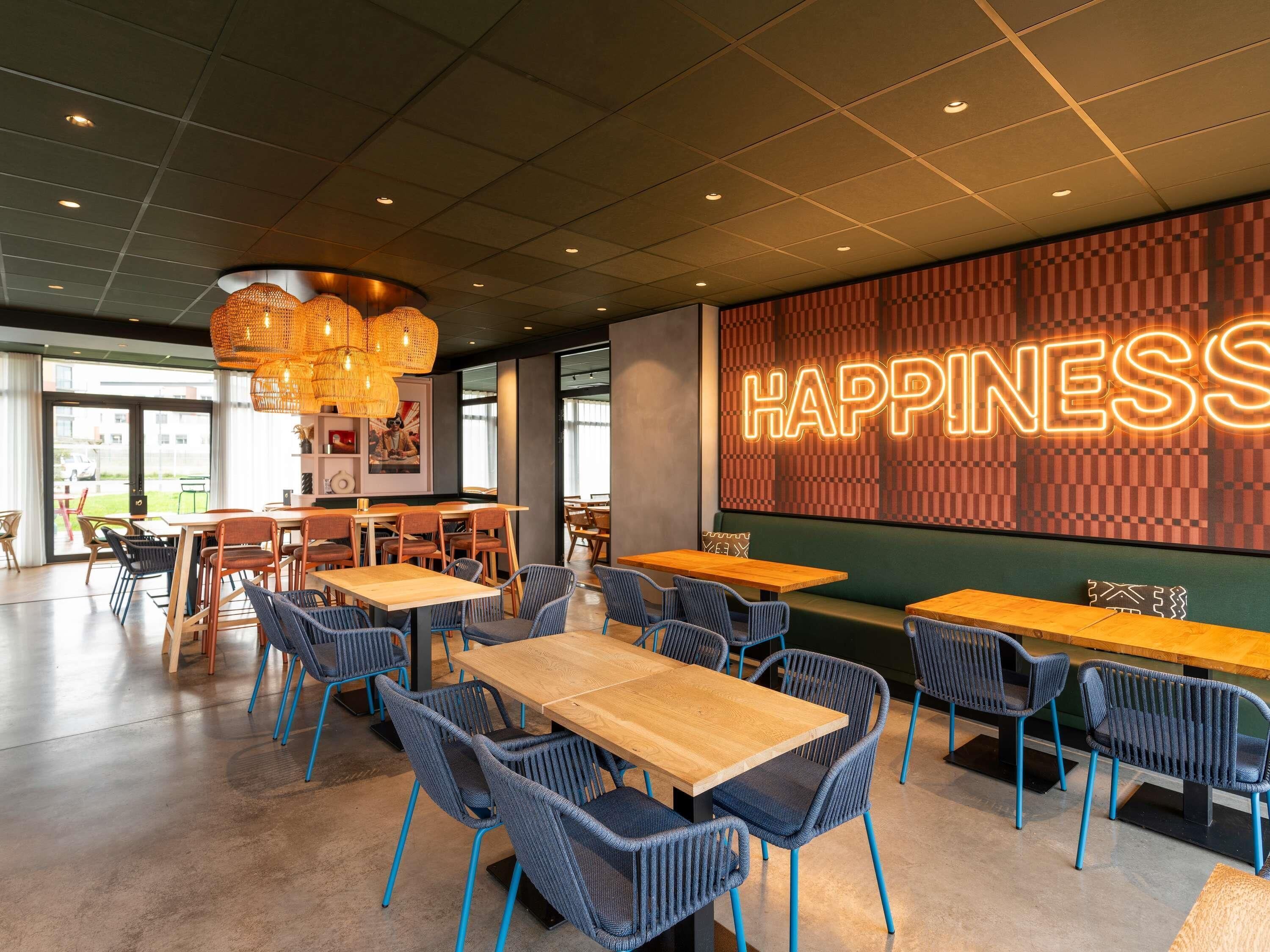 A modern restaurant interior with wooden tables and blue chairs, a neon 'HAPPINESS' sign, and a patterned wall.