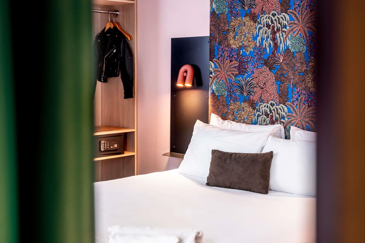 A hotel room with a bed, a wardrobe, and a wall with a colorful floral pattern.
