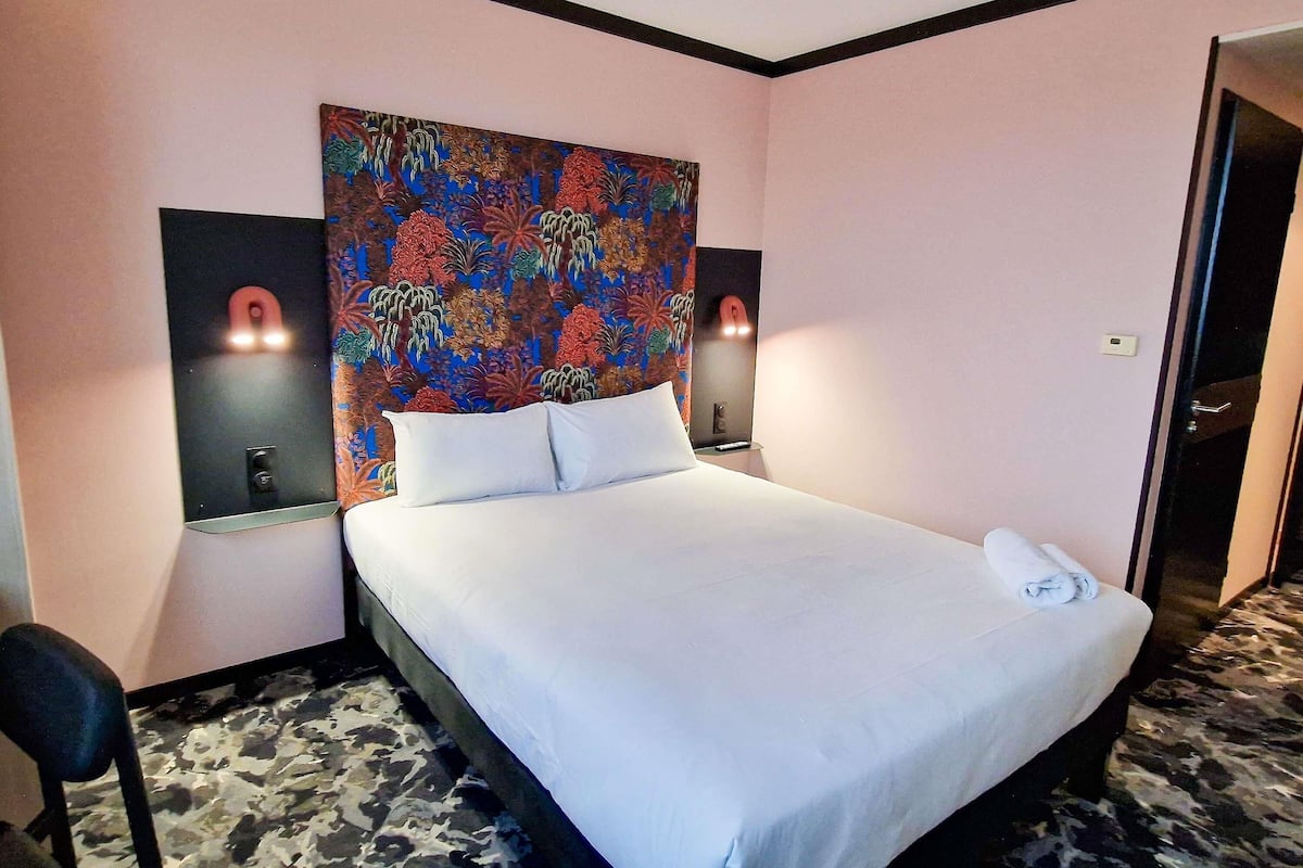 A hotel room with a bed, two wall-mounted lamps, a colorful floral headboard, and a door.