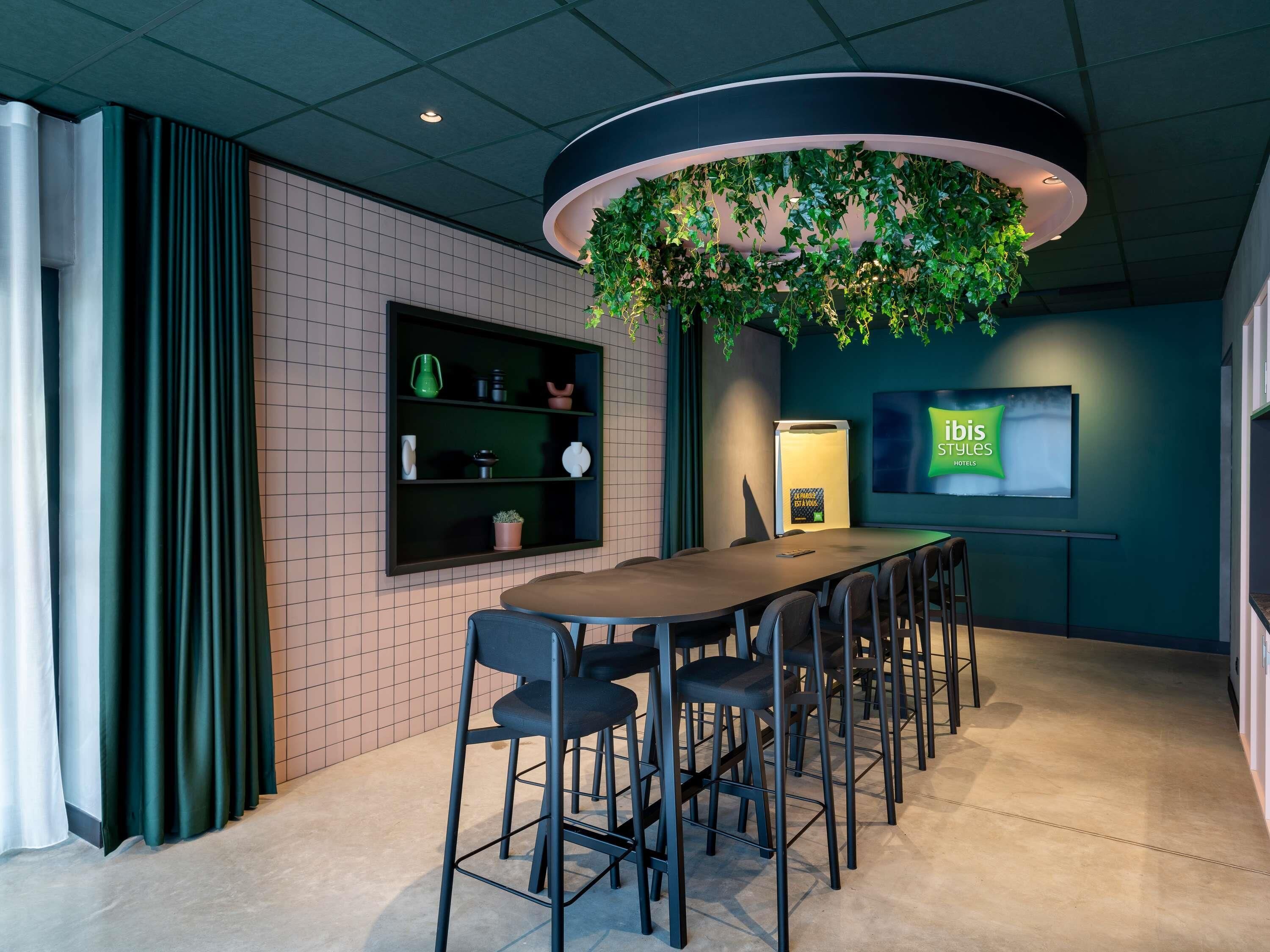 A conference room with a long table, chairs, a shelf with items, a TV screen, and a green plant hanging from the ceiling.