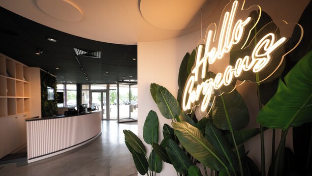 Een moderne receptie met een neonbord waarop 'Hello Guests' staat, grote groene planten en een gebogen receptiebalie.