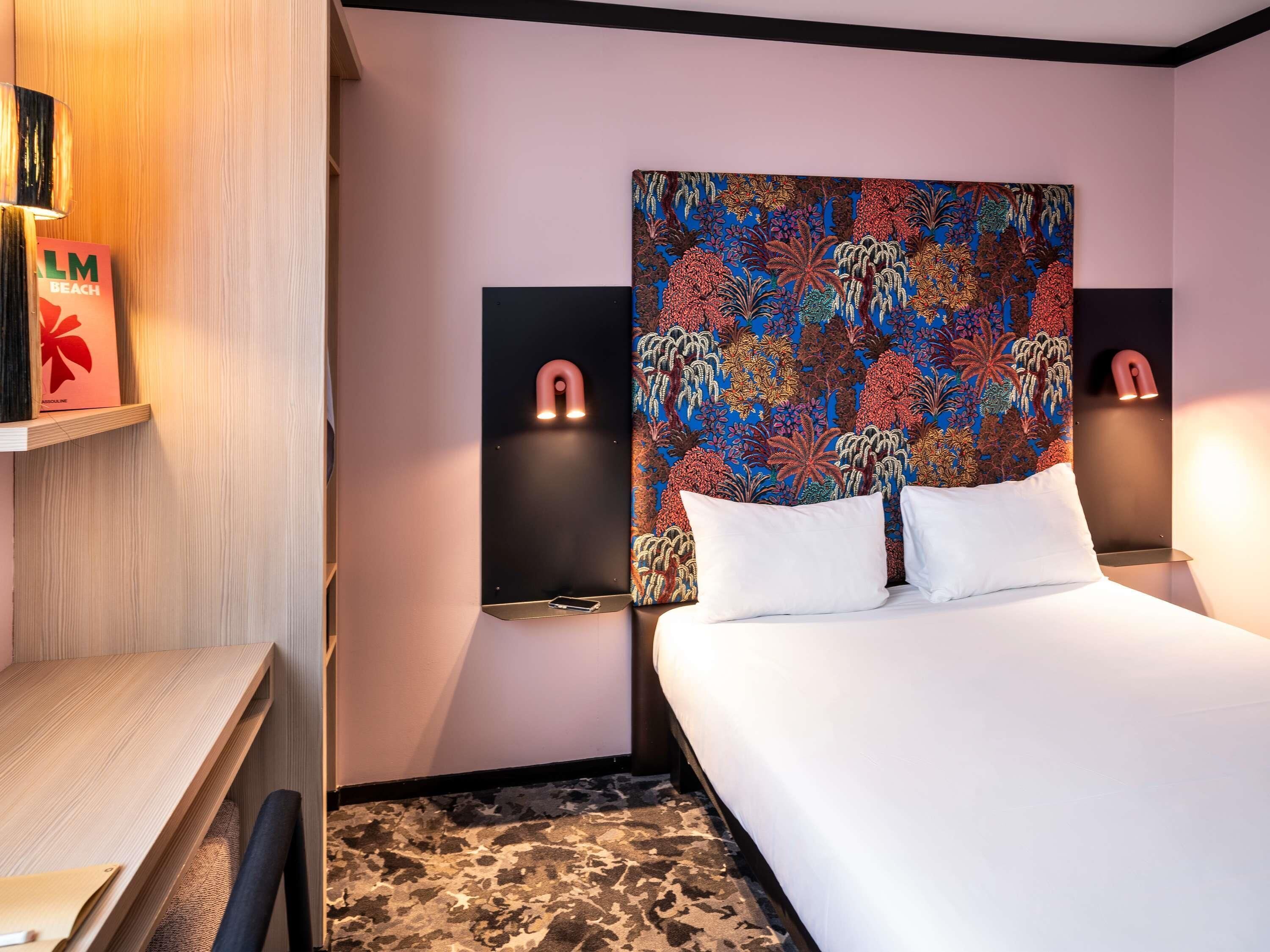 Habitación de hotel con cama, escritorio, silla, cabecera estampada y dos lámparas de pared.