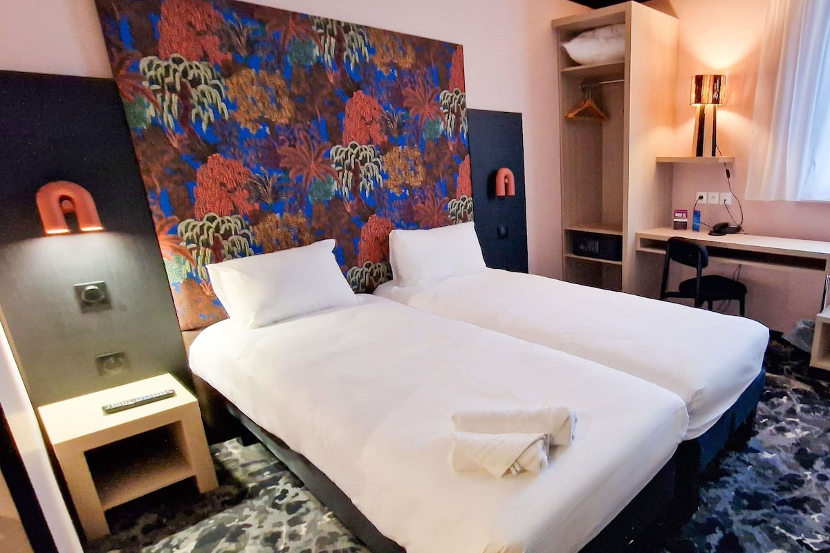 A hotel room with two beds, a desk, a chair, a wardrobe, and a colorful abstract painting on the wall.