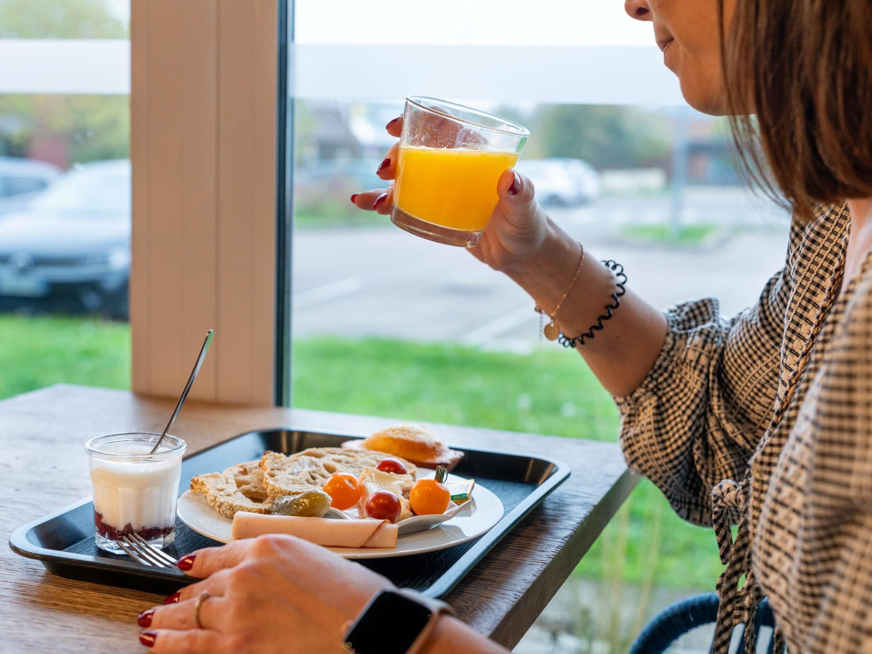 En person som håller i ett glas apelsinjuice och sitter vid ett bord med en bricka framför sig. På brickan finns en smörgås, ost, oliver och cocktailtomater.
