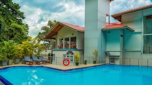 Una piscina con sillas de descanso y un flotador salvavidas, rodeada de exuberante vegetación y un edificio con tejado rojo.