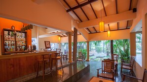 A bar area with wooden stools, a bar counter with liquor bottles, and a view of a tropical outdoor area with chairs and tables. - Pelwehera Village Resort (Dambulla)