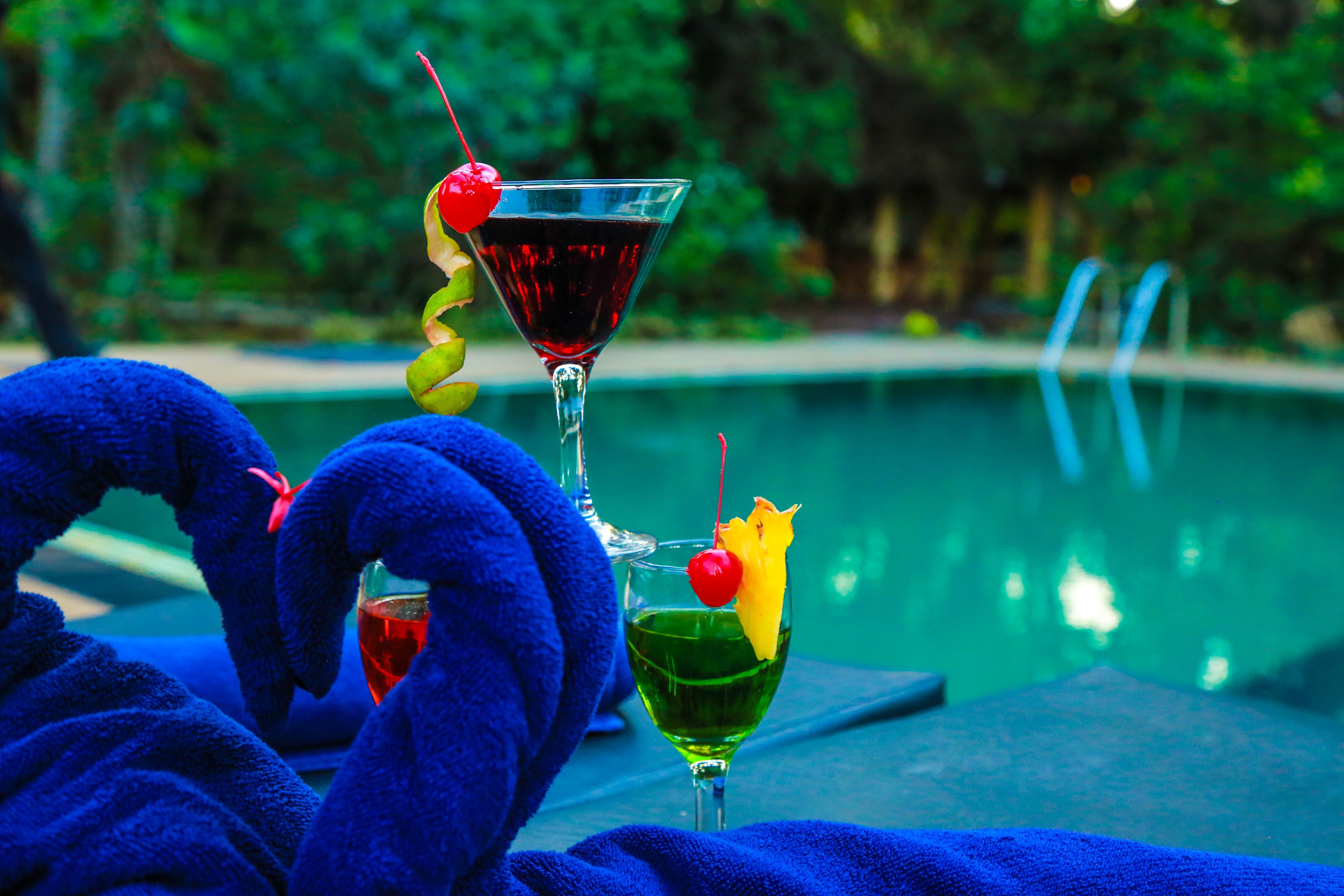 A poolside setting with two drinks, one in a martini glass and another in a cocktail glass, each garnished with a cherry and a slice of fruit.