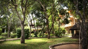 A lush courtyard with mature trees, a thatched-roof structure, and a building with balconies in the background. - Pelwehera Village Resort (Dambulla)