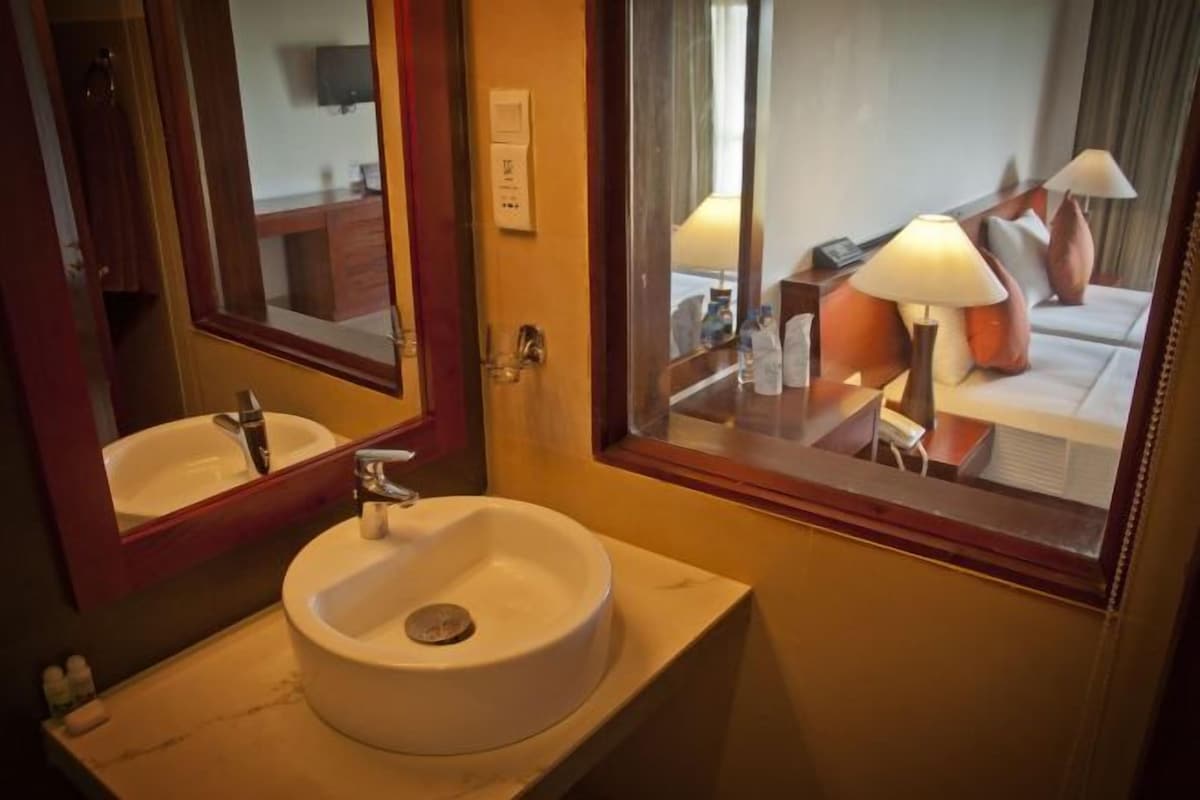 Une salle de bain d’hôtel avec un lavabo, un miroir et une fenêtre donnant sur un lit et une lampe.