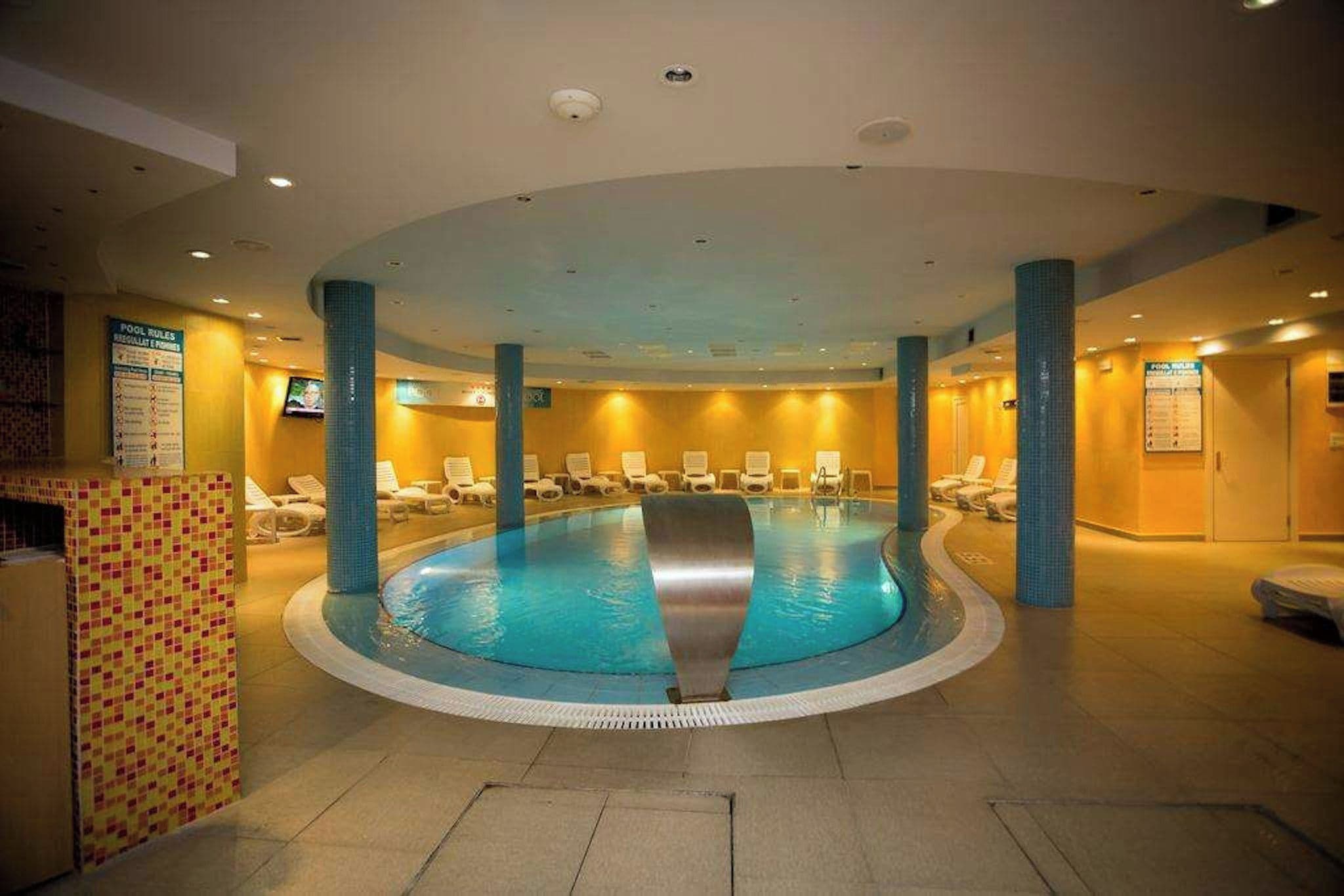Una amplia piscina interior con un tobogán central, rodeada de sillones de descanso y un suelo de baldosas.