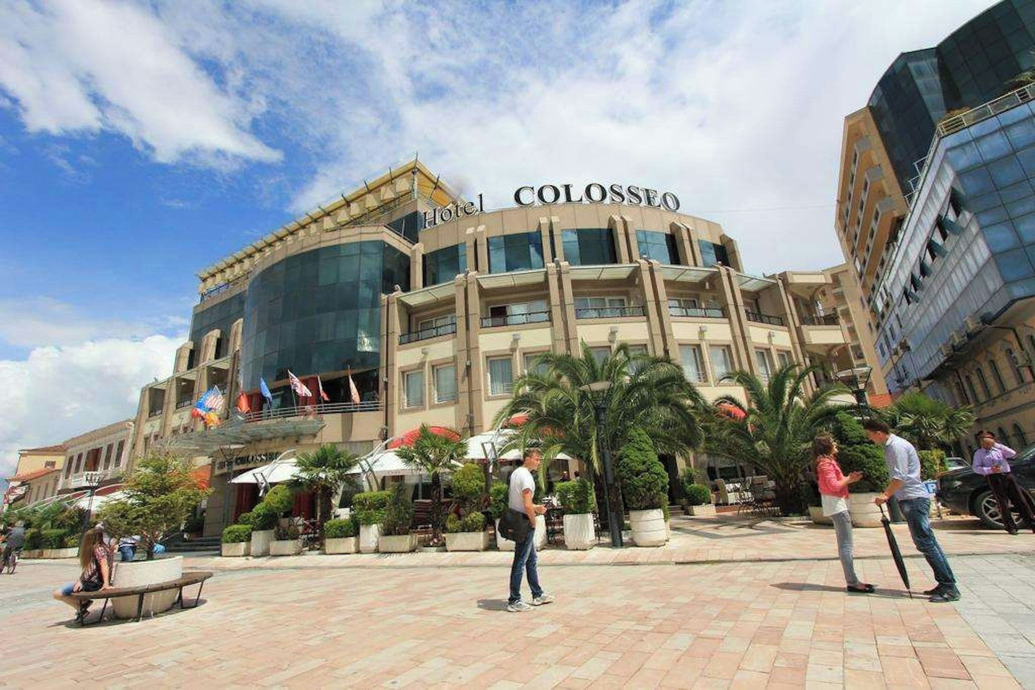 Un hotel con fachada moderna, varios balcones y un patio central con palmeras y mobiliario exterior.