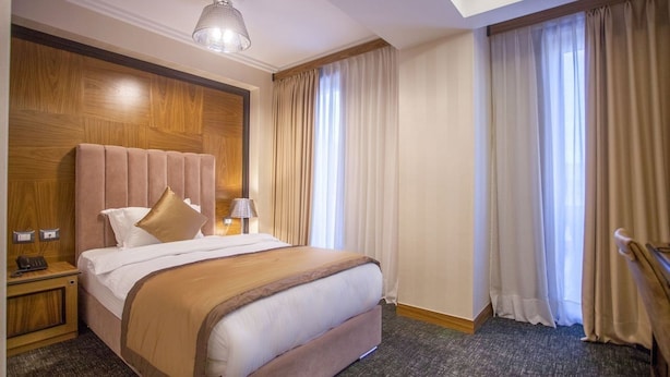 A hotel room with a large bed, wooden headboard, a nightstand with a lamp, and a view of the outside through large windows with curtains.