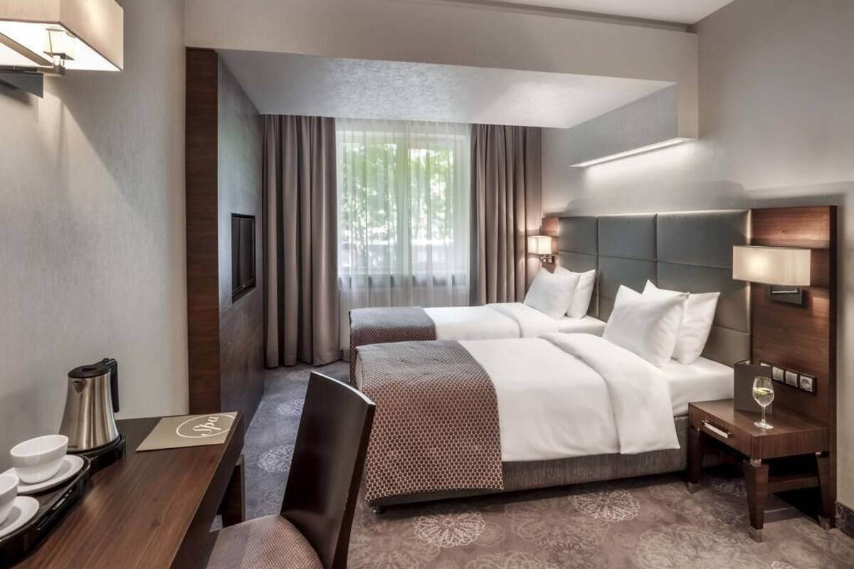 Une chambre d’hôtel avec deux lits, un bureau, une chaise, une table de chevet avec une lampe et une fenêtre avec des rideaux.