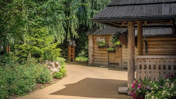 A wooden cabin with a thatched roof, surrounded by lush greenery and a paved pathway.