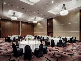 A banquet hall with round tables, black chairs, and chandeliers.