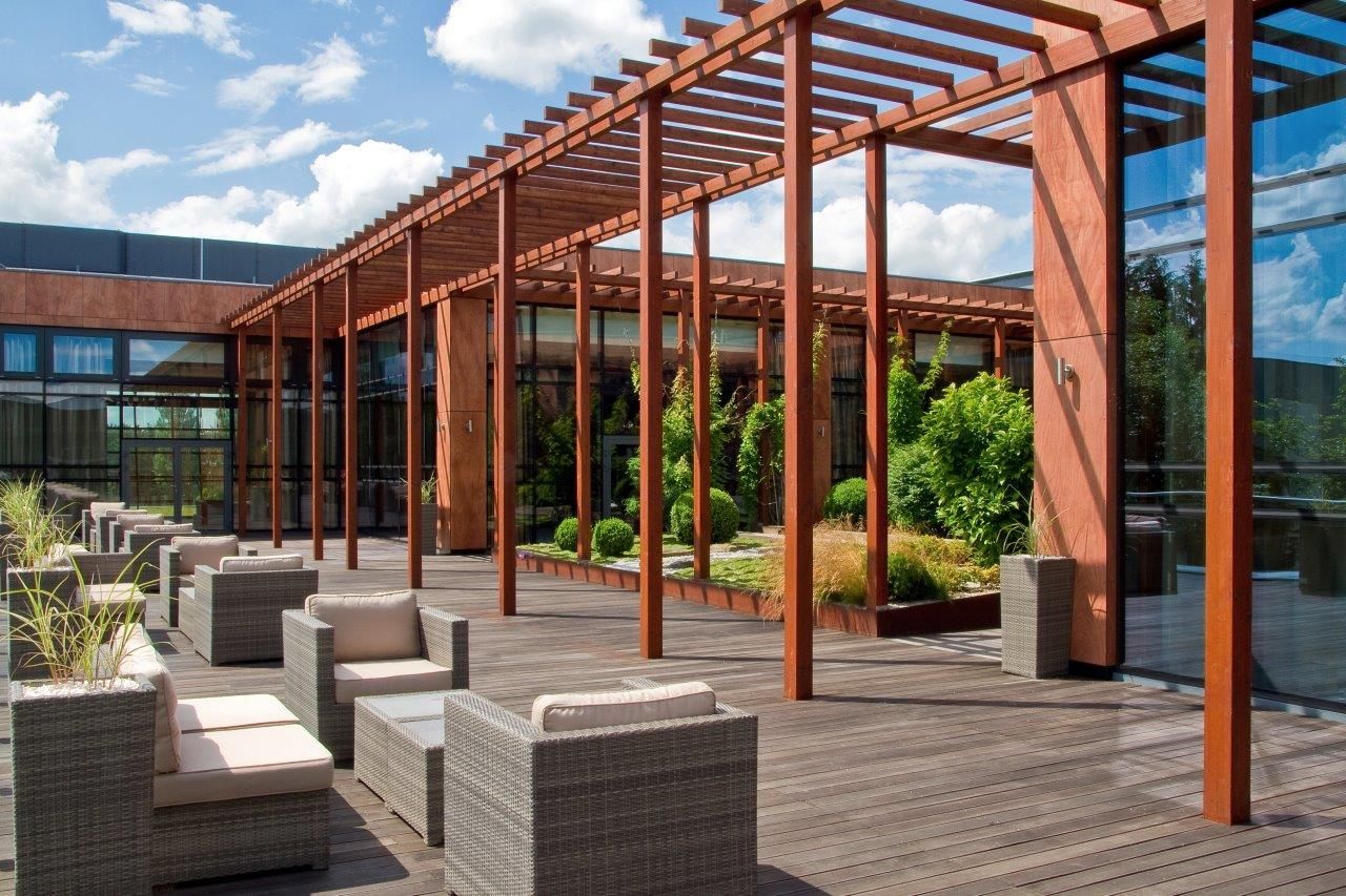 Un espacio al aire libre techado con pérgolas de madera, asientos y vistas a zonas verdes y un edificio.