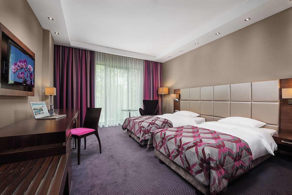 Hotellihuone, jossa on kaksi sänkyä, työpöytä kannettavalla tietokoneella, tuoli ja seinälle asennettu televisio.