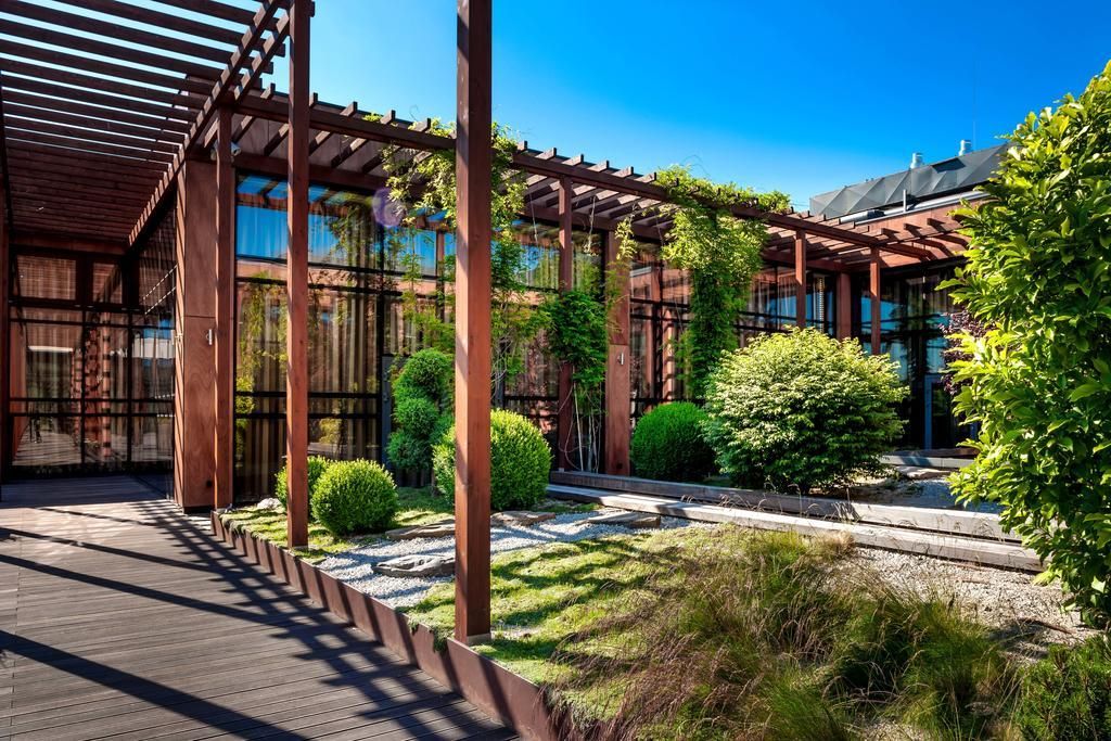 Ein modernes Gebäude mit einer Holzpergola, Glasfassaden und einem Garten mit Sträuchern und einem Weg.