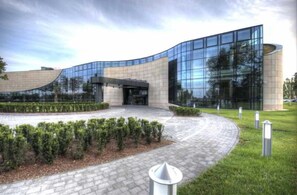 A modern building with a curved glass facade, a paved entrance area, and neatly trimmed hedges. - Hotel Warszawianka (Serock)