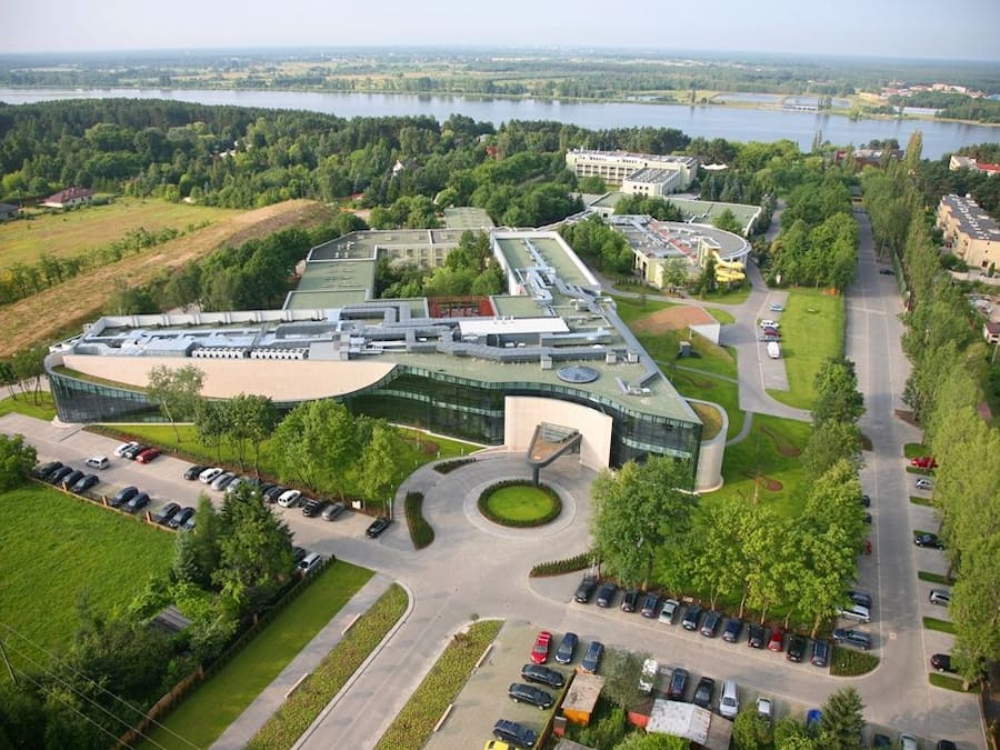 A modern building with a curved roof, surrounded by parking and greenery, overlooking a body of water.