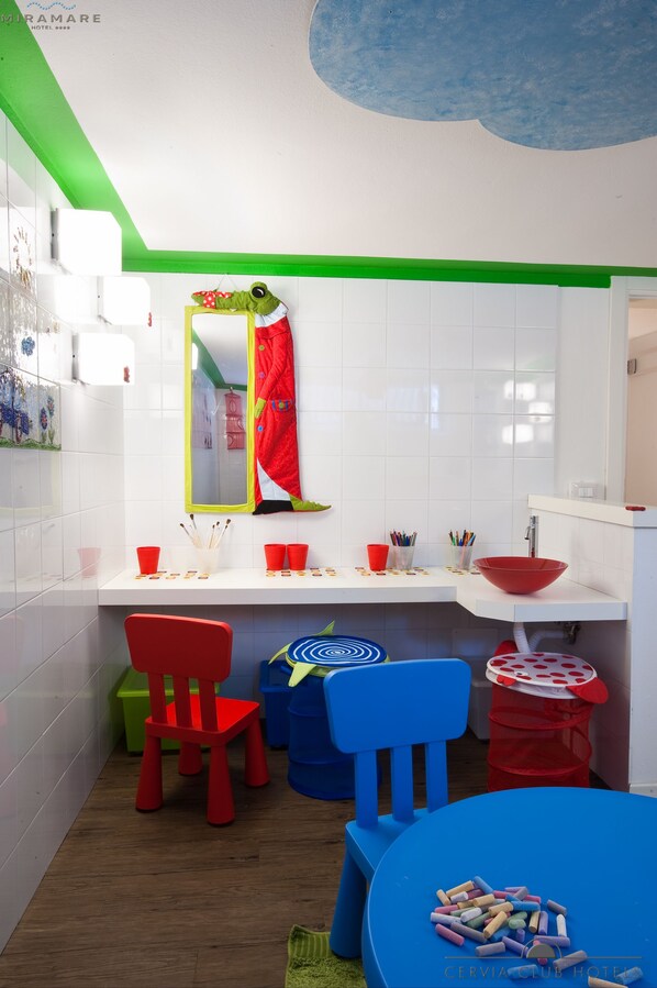 A children's room with a white tiled wall, a green ceiling, a red chair, a blue table, and a red trash bin. - Hotel Miramare (Cervia)