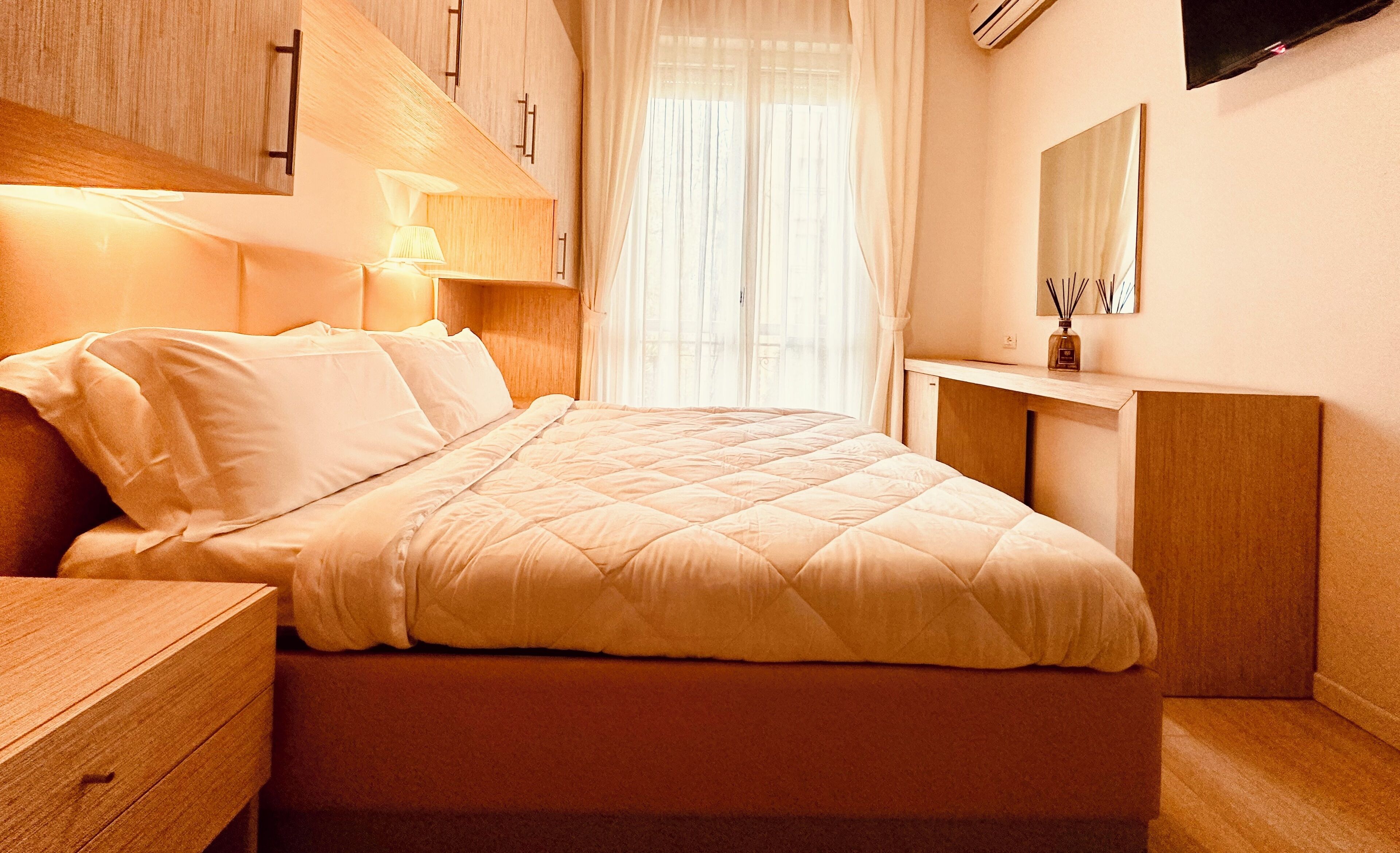 A neatly made bed with a quilted cover, a bedside table, a lamp, a mirror, and a window with sheer curtains.