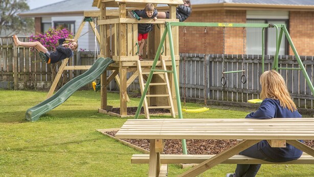 A wooden play structure with a slide and climbing area, a picnic table, and a fenced yard.