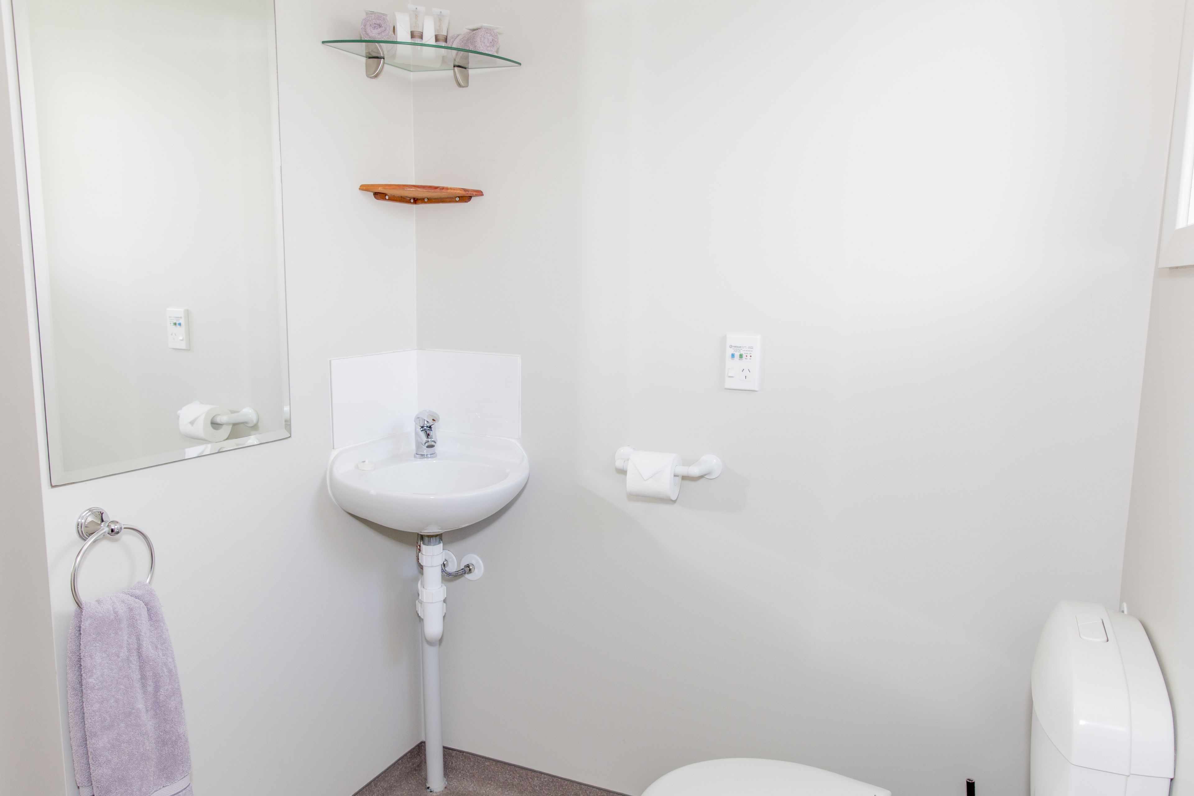 Un baño con lavamanos, espejo y retrete blancos. Se aprecia un sujetapapeles con una toalla.