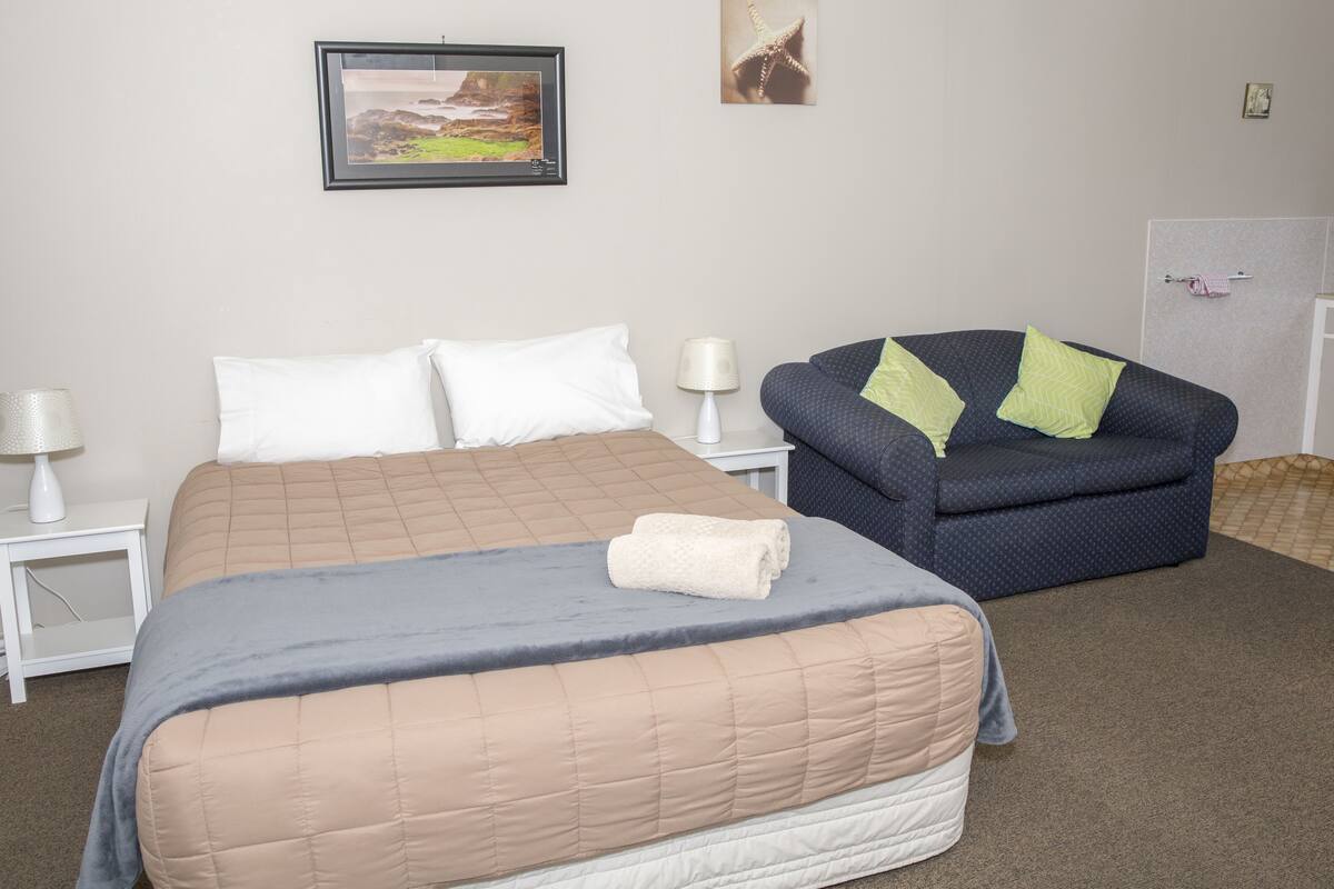 Un dormitorio con cama, una silla, una mesita de noche, una lámpara y un cuadro en la pared.