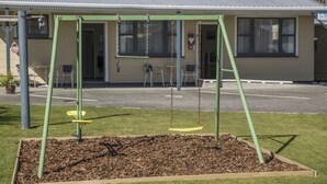 A playground with swings and a yellow seat, surrounded by mulch and a wooden border, in front of a building with a metal roof and windows.