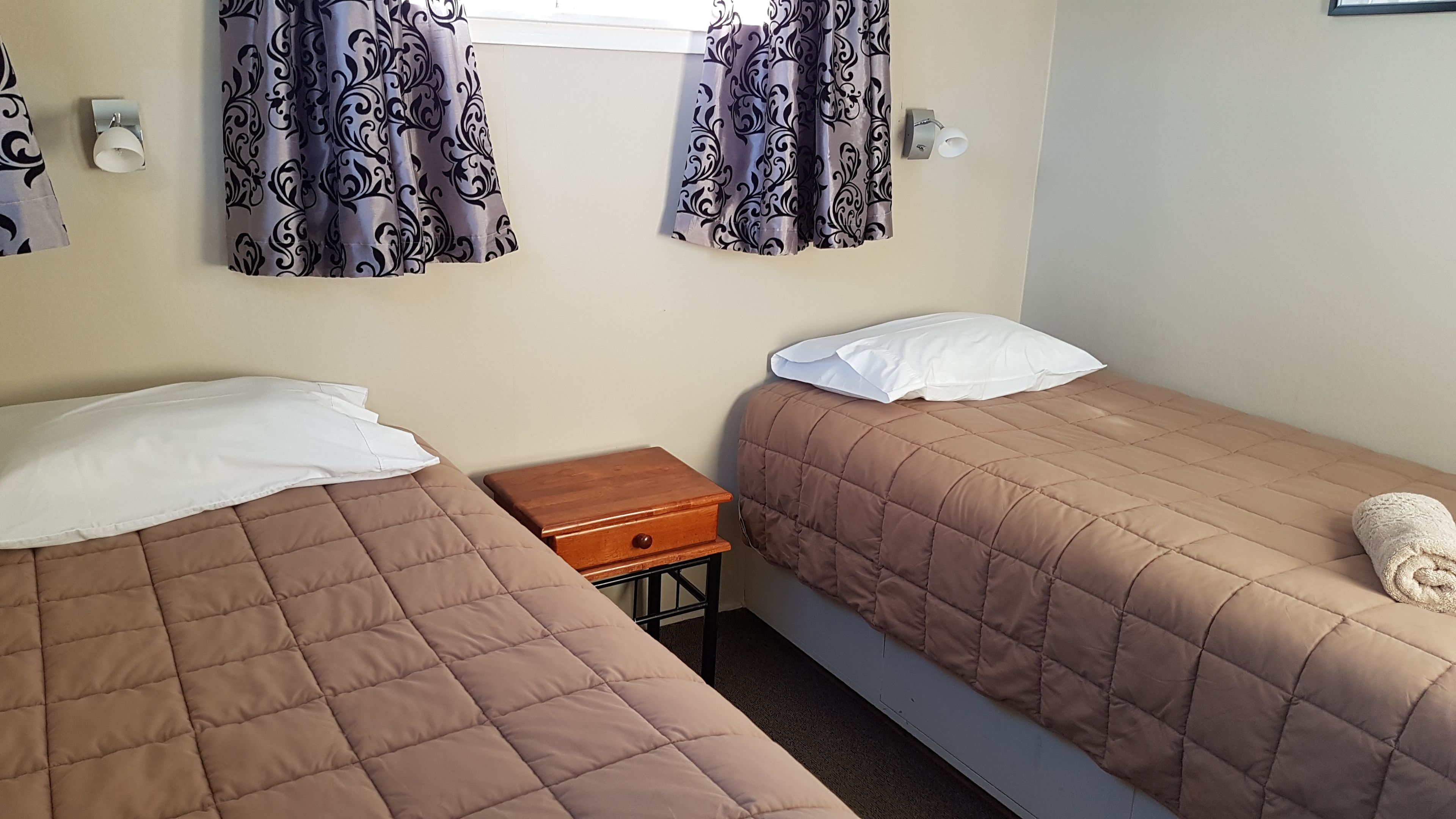 A room with two beds, each with a brown quilted cover and white pillows. There is a small wooden nightstand with a drawer between the beds.