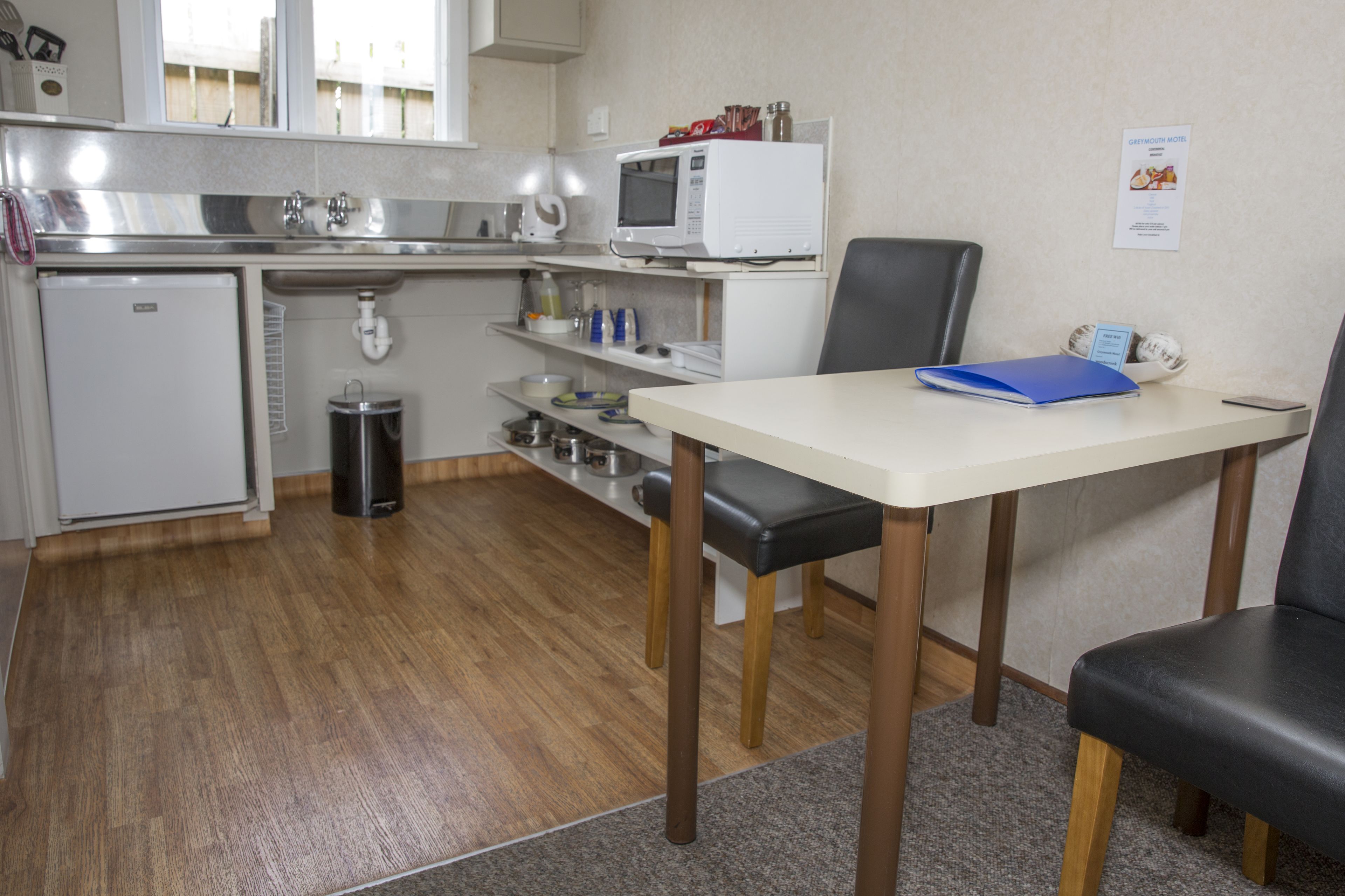 A compact kitchen with a refrigerator, microwave, sink, and a small trash bin. There is a table with a blue folder and a chair.