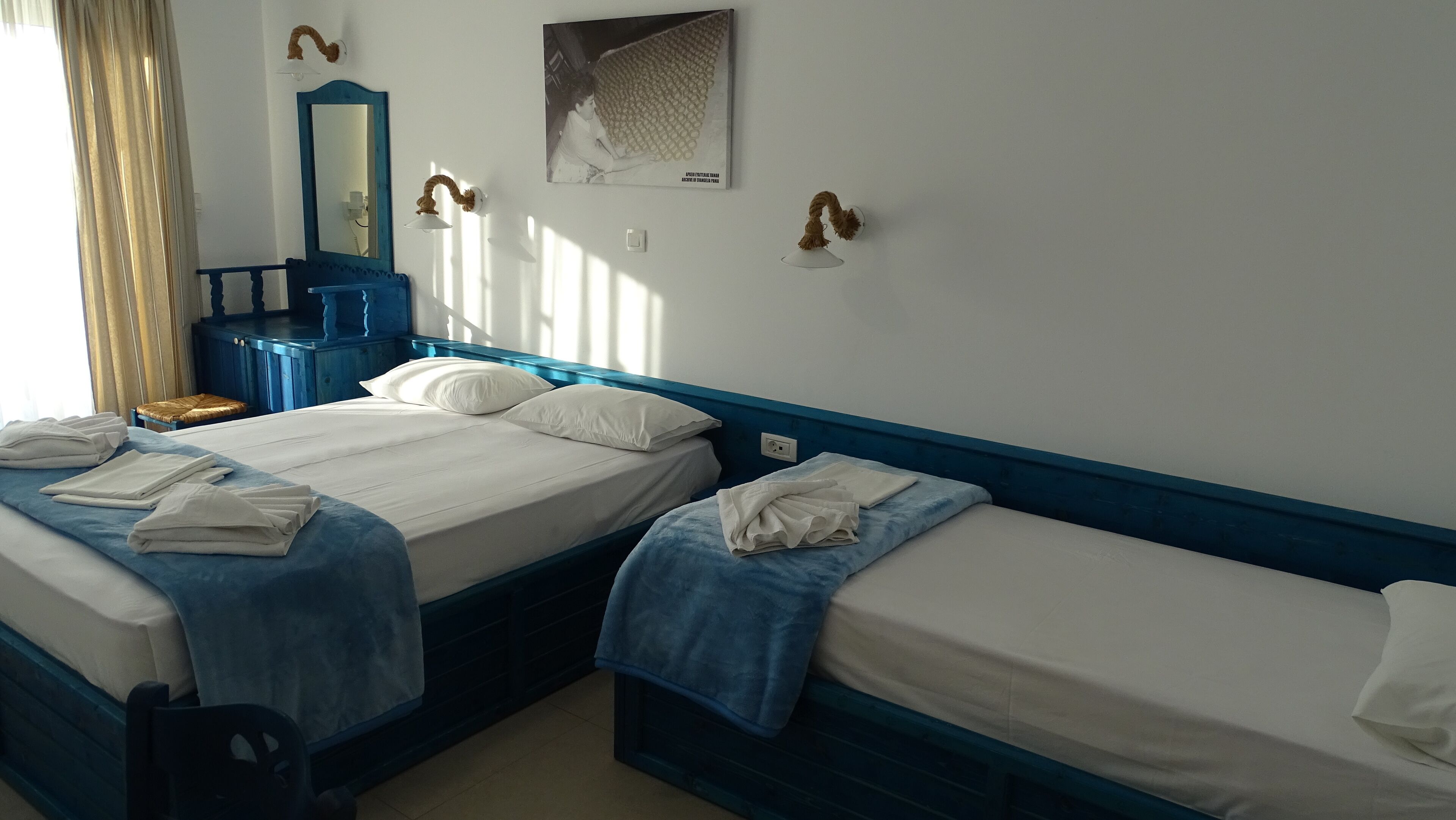 A hotel room with two beds, a blue headboard, a mirror, a painting, and a window with curtains.