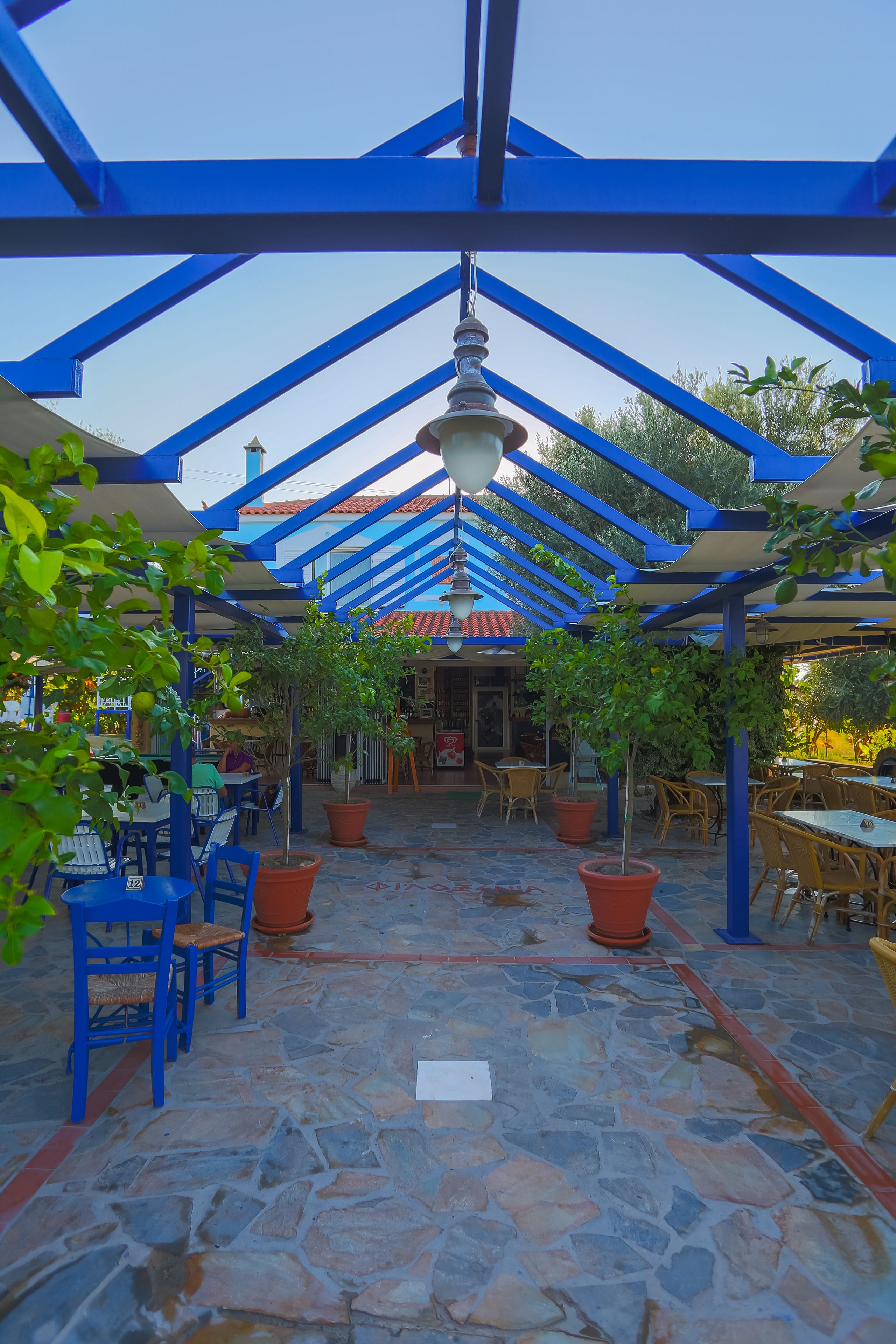 Een terras buiten met stenen vloer, blauwe metalen balken en plantenbakken.