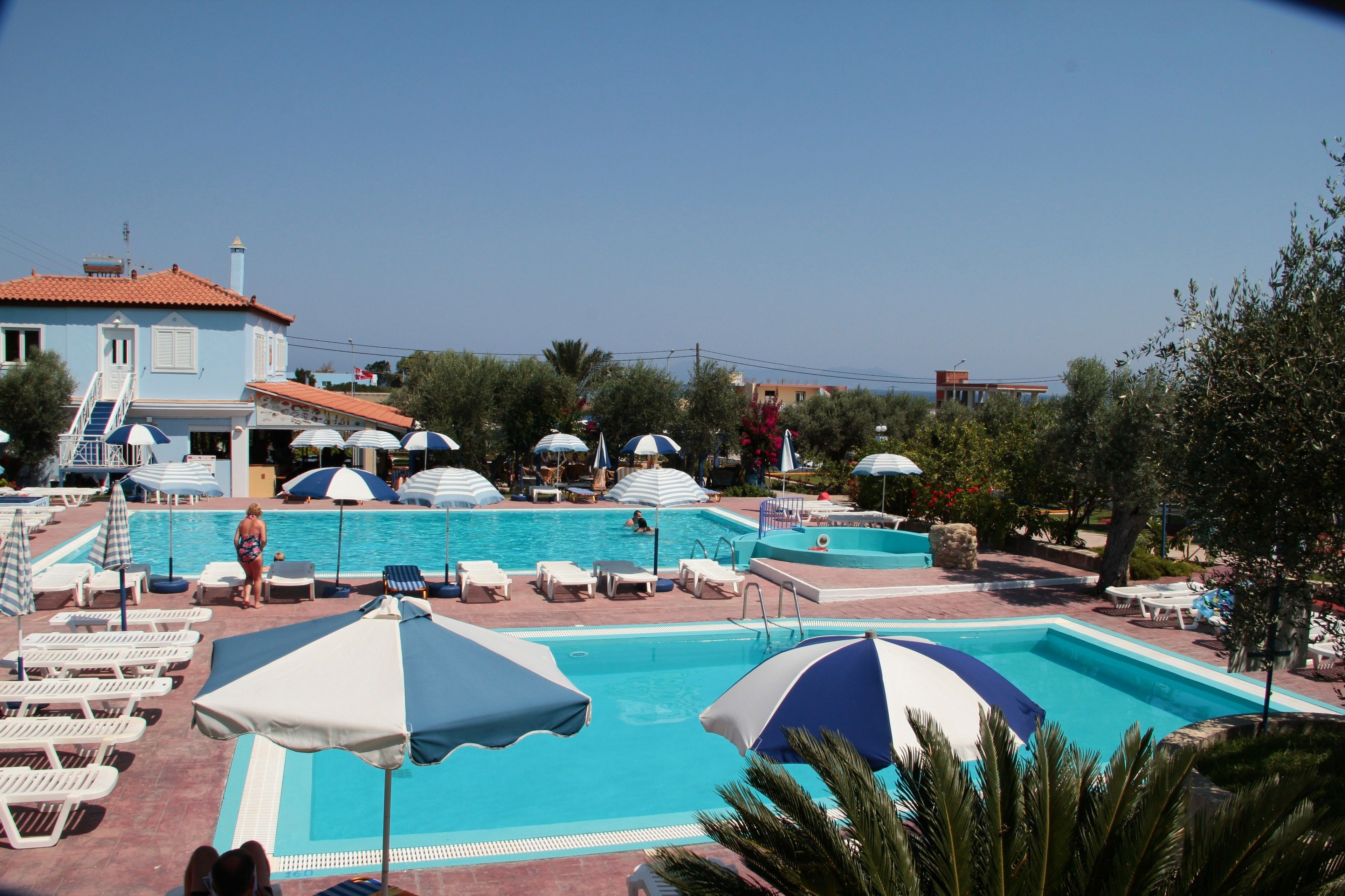 A swimming pool with lounge chairs and umbrellas, surrounded by trees and a building with a red roof.