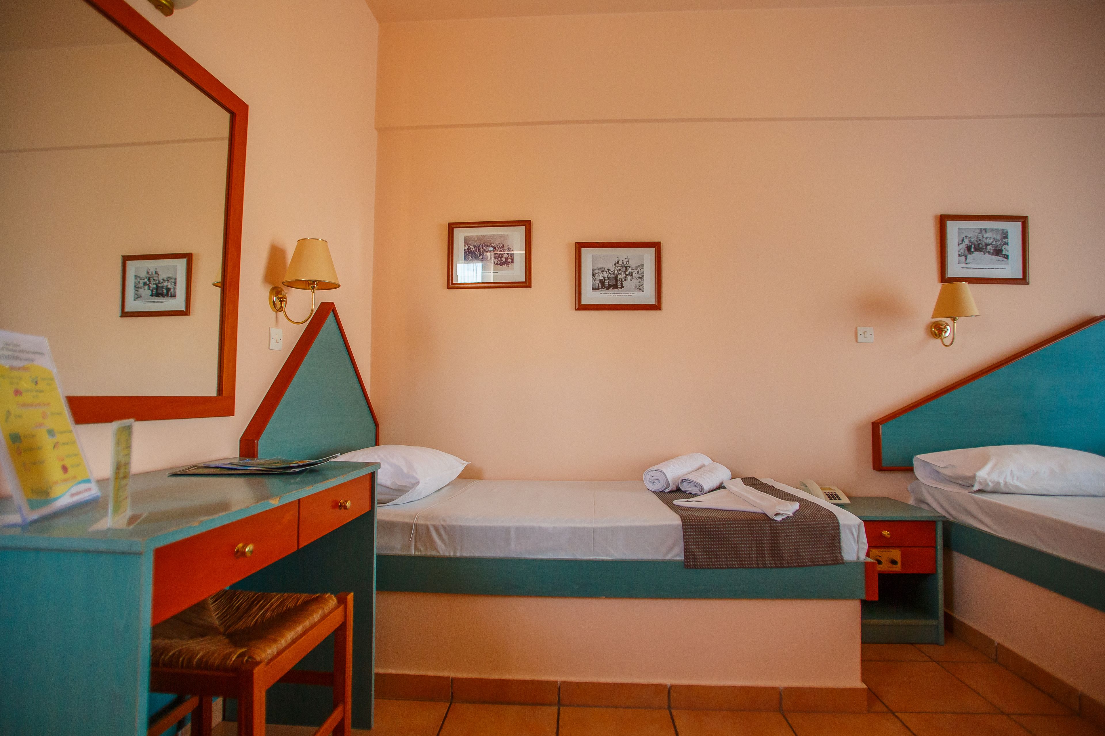 Habitación de hotel con dos camas individuales, un escritorio con silla, un espejo y tres cuadros en la pared.