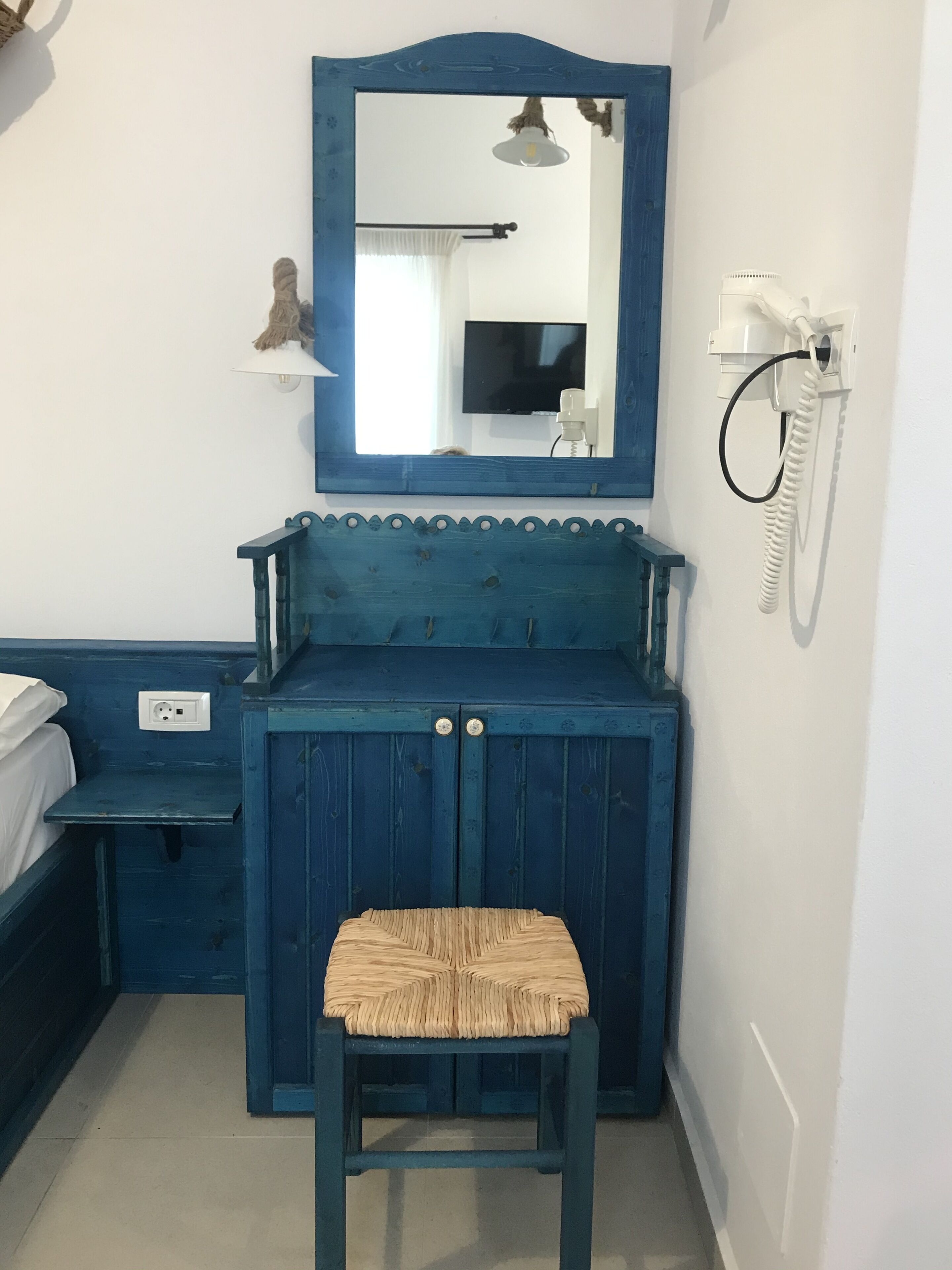 A small, blue-painted room with a wooden cabinet, a chair, a bed, a mirror, a wall-mounted phone, and a television.