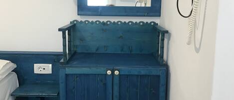 A small, blue-painted room with a wooden cabinet, a chair, a bed, a mirror, a wall-mounted phone, and a television.