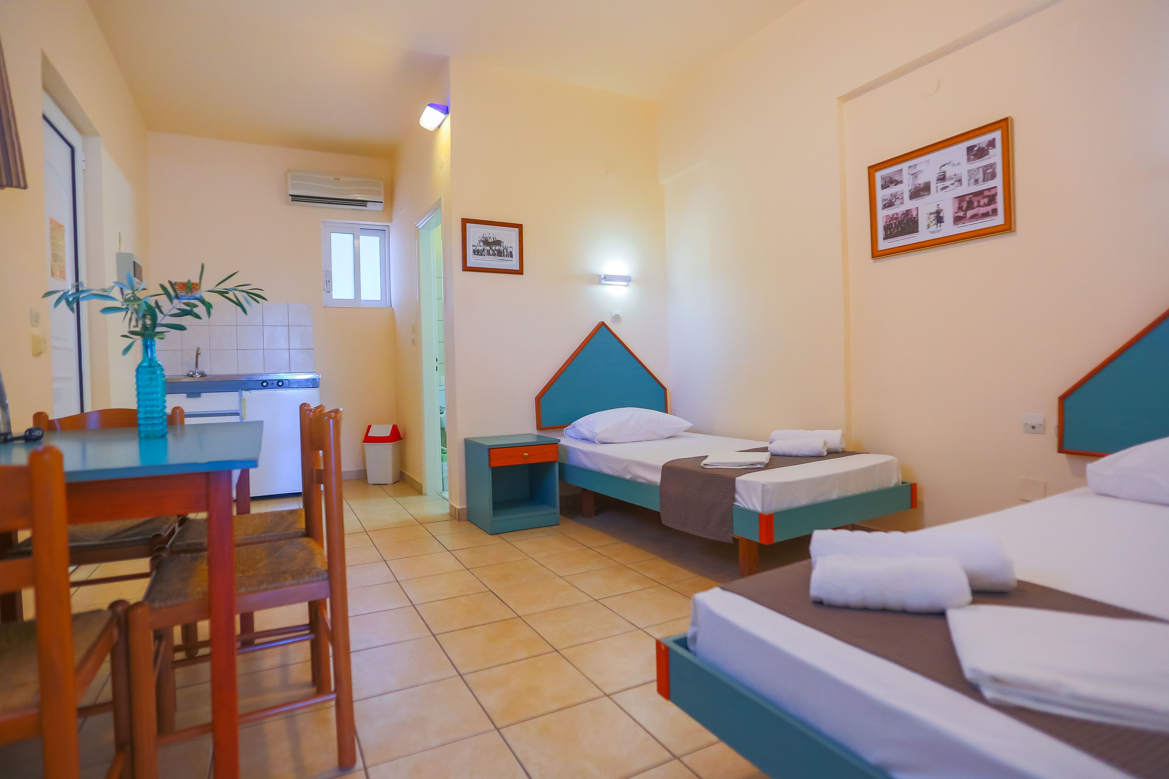 Une chambre d’hôtel avec deux lits simples, une petite kitchenette, une table à manger avec des chaises et une plante en pot.