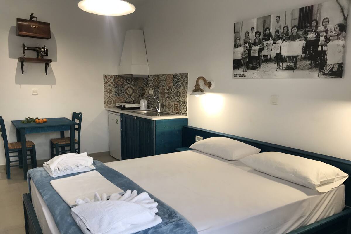 A bedroom with a bed, pillows, a bedside table, a desk, a chair, a small kitchenette, and a wall-mounted shelf.