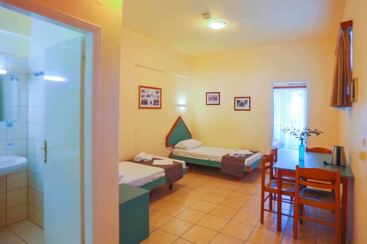 A room with two single beds, a small table with chairs, a sink, and framed pictures on the wall.