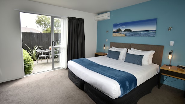 A bedroom with a large bed, a sliding glass door to a patio, a painting of a beach scene, and a nightstand with a lamp. - Fyffe On Riccarton (Christchurch)