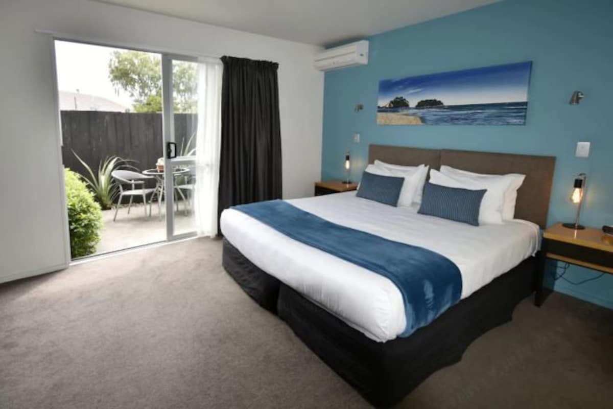 A bedroom with a large bed, a sliding glass door to a patio, a painting on the wall, and a nightstand with a lamp.