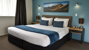 A large bed with a blue and white bedspread, two bedside tables with lamps, a painting on the wall, and a window with curtains. - Fyffe On Riccarton (Christchurch)