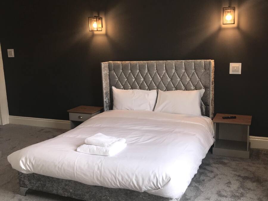 Un dormitorio con cabecera capitonada, ropa de cama blanca, dos mesitas de noche y un control remoto sobre una de ellas.
