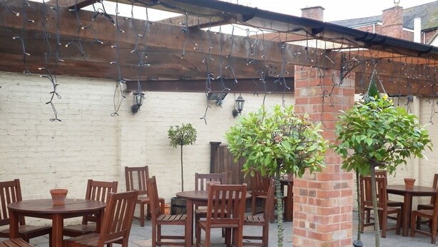 An outdoor patio with wooden tables and chairs, potted plants, and a brick wall. - Dixie Arms (Nuneaton)