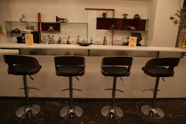 Zona de bar con cuatro taburetes, una barra con diferentes botellas y vasos, y objetos decorativos en estantes.