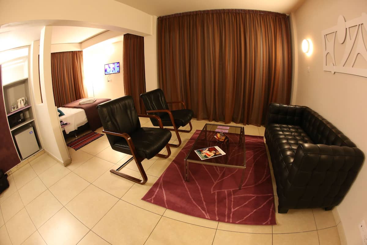 Habitación de hotel con una cama, dos sillones de cuero negro, una mesa de centro de vidrio, una alfombra roja, un sofá de cuero negro y un televisor en la pared.