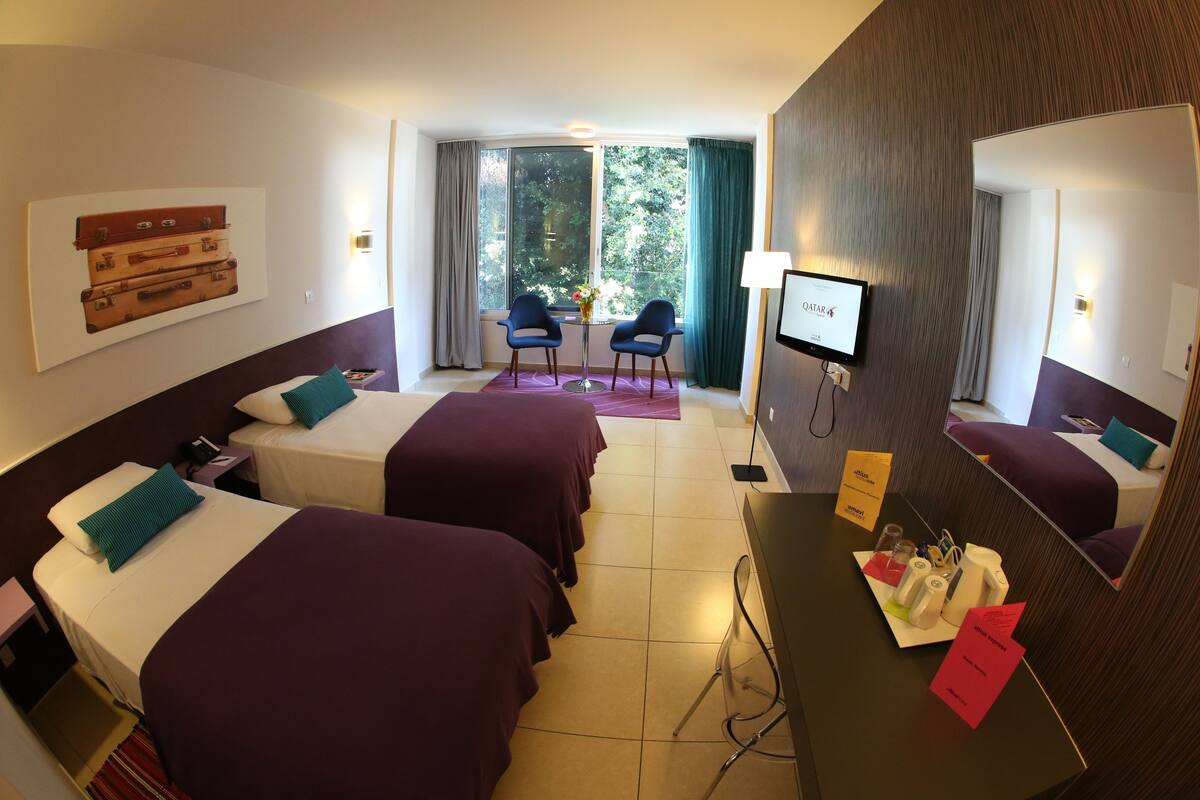 Una habitación de hotel con dos camas, un escritorio con espejo, un televisor y una ventana con cortinas.