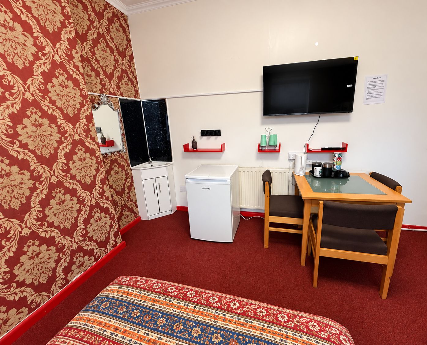 A small, furnished room with a bed, a desk, a chair, a television, a radiator, and a wardrobe.